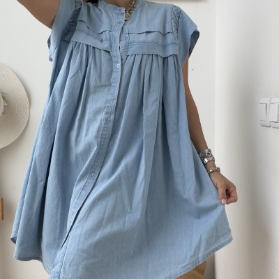Vestido azul claro estilo solto com botões frontais e pregas, mulher usando óculos, acessórios, ambiente interior com parede branca.