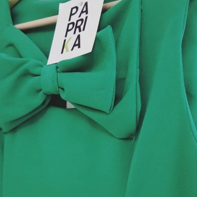 Vestido verde com laço e etiqueta PAPRIKA pendurado em cabide.
