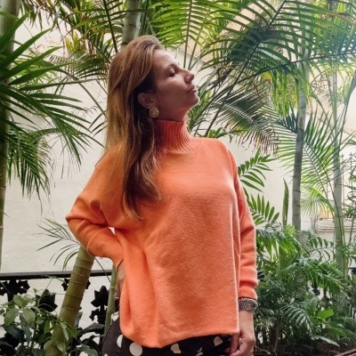 Mulher com camisola tangerina de malha e calças pretas com bolinhas brancas num ambiente com plantas tropicais.