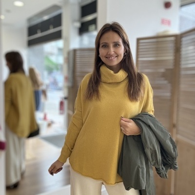 Mulher com camisola amarela, calças claras e casaco verde num ambiente interior moderno