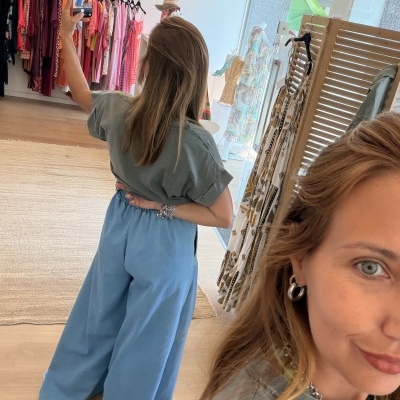 Mulher de costas vestindo t-shirt cinza claro e calças azuis largas em loja de roupa com vestidos coloridos ao fundo.