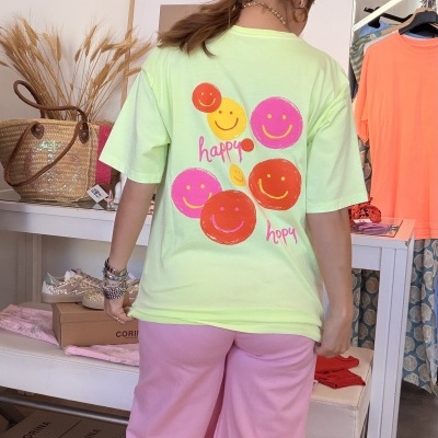 T-shirt verde claro com carinhas sorridentes coloridas e texto happy hopy na parte de trás, calças largas rosas e sapatilhas com brilho.