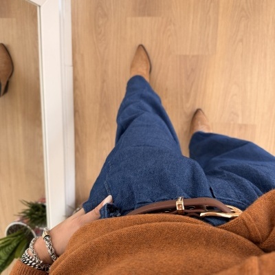 Roupa com calças de ganga azul, suéter castanho, cinto castanho, botas castanhas e pulseiras prateadas no pulso, vista de cima para baixo.