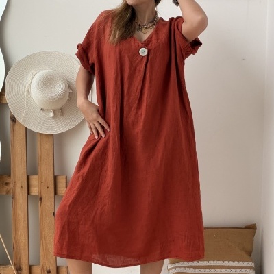 Mulher a usar vestido midi terracota com botão branco e acessórios, chapéu branco pendurado em suporte de madeira e quadro branco na parede