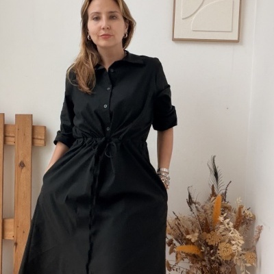 Mulher com vestido preto e botas pretas em ambiente interior com vaso de flores secas