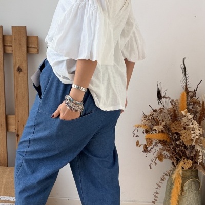 Mulher com blusa branca de mangas bufantes, calças azuis largas e sapatilhas douradas em sala com decoração simples