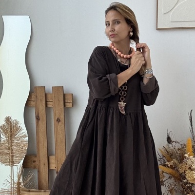 Mulher com vestido castanho-escuro comprido e colares, em ambiente interior com decoração neutra.