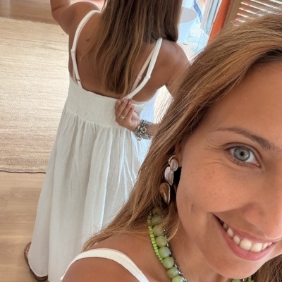 Mulher com vestido branco e colares verdes sorrindo em ambiente interno com roupas ao fundo