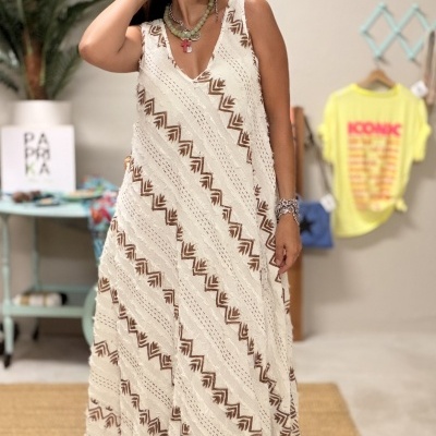 Mulher com vestido longo branco e castanho com padrão geométrico, sandálias plataforma pretas e colares, em ambiente interior decorado.