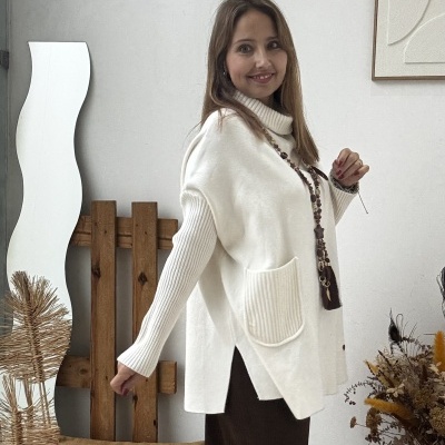 Mulher com camisola branca de manga comprida e gola alta junto a almofada castanha e elementos decorativos numa sala interior.