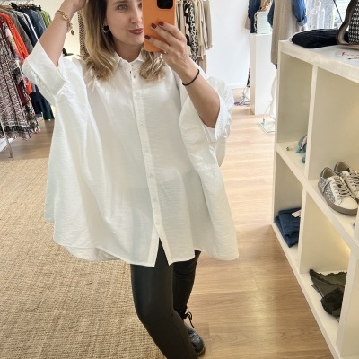 Mulher com blusa branca larga e calças pretas a tirar selfie num espelho numa loja de roupas