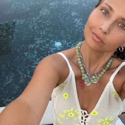 Top branco com bordados amarelos fluorescentes e colar de contas verdes e correntes metálicas num ambiente com piscina ao fundo