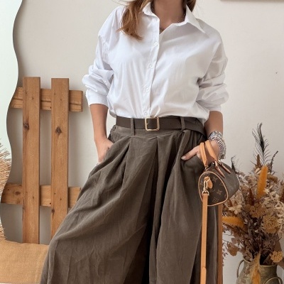 Mulher com camisa branca e calças largas verdes em ambiente interior com decoração rústica