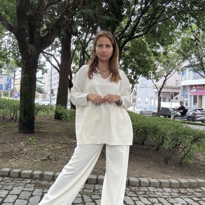 Mulher com conjunto branco de malha, suéter gola V e calças largas, em passeio urbano com árvores e calçada de pedra