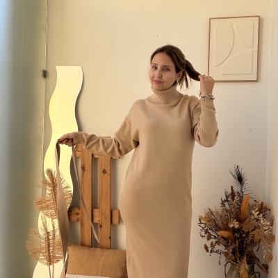 Mulher com vestido comprido bege de malha e gola alta num interior com decoração neutra e elementos naturais