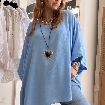Mulher com blusa oversized azul claro, calças de ganga, colar com pendente, pulseiras e óculos num interior com roupas ao fundo.