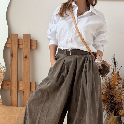 Mulher com camisa branca e calças largas verdes com cinto castanho e bolsa monograma num ambiente interior rústico