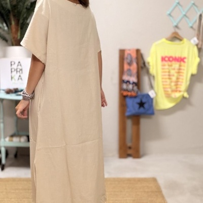 Mulher de vestido comprido bege num ambiente interior com planta, tapete e prateleira azul