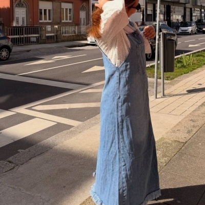 Vestido de ganga azul claro com bainha desfiada, blusa branca texturada e sneakers creme com dourado numa rua urbana