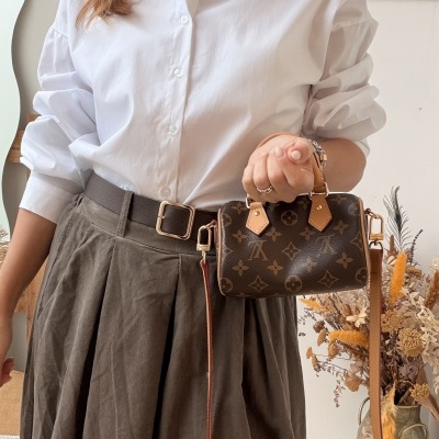 Mulher com camisa branca e saia castanha segurando pequena bolsa castanha com correia longa numa sala