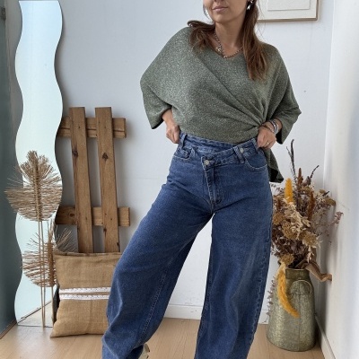 Mulher vestindo calças de ganga largas azuis e camisola verde num interior com espelho e flores secas