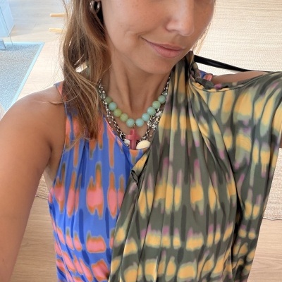Mulher com colares e brincos vestindo roupa colorida e segurando outra peça colorida.