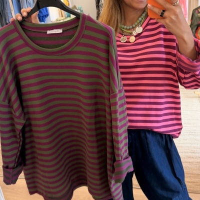 Camisola às riscas verde e bordeaux num cabide e mulher a usar camisola às riscas rosa, num ambiente de loja de roupa