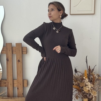 Mulher com vestido castanho escuro de malha pregueada e botas pretas em ambiente interior minimalista