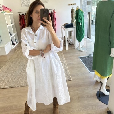 Vestido branco comprido de tecido leve com botões castanhos e gola de camisa, em loja de roupa com manequins e chão de madeira clara