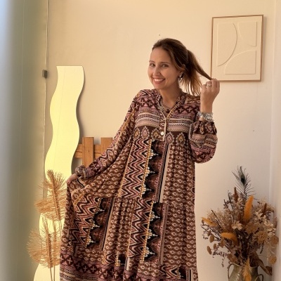 Mulher com vestido comprido padrão étnico numa sala clara com plantas secas e espelho