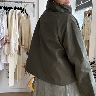 Casaco curto verde-oliva com gola subida e vestido cinzento comprido usado por pessoa em ambiente interior de loja.