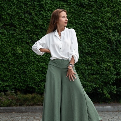 Mulher com calças largas verdes e camisa branca de mangas arregaçadas em pavimento de pedra com fundo de plantas.