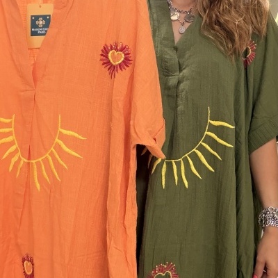 Vestidos de algodão largo, laranja e verde, com bordados de sol e coração, apresentados por mulher com cabelo loiro