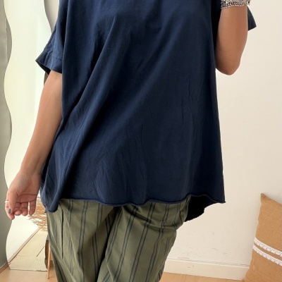 Mulher com t-shirt azul escura e calças verde-oliva riscas, bijutaria prateada e colar colorido, cenário interior com chão de madeira