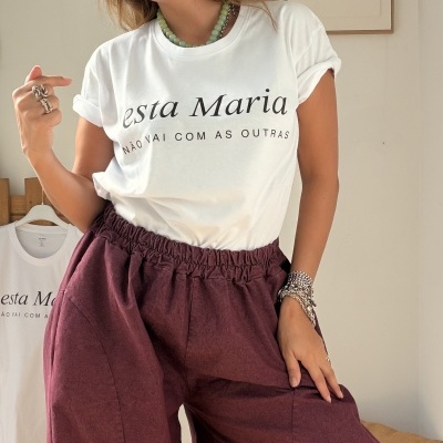 Mulher a usar t-shirt branca com texto preto e calças largas castanho avermelhadas, em ambiente interior