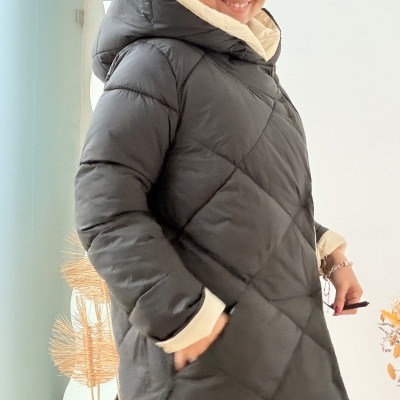 Casaco acolchoado comprido preto com capuz bege e bolsos, mulher sorrindo num espaço interior com decoração seca