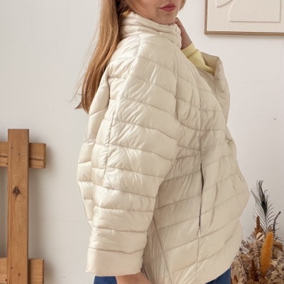 Casaco acolchoado creme com padrão horizontal em mulher com calças azuis, fundo branco e decoração natural