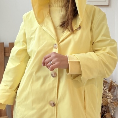 Mulher com casaco amarelo com capuz e botões, camisola de gola alta amarela, jeans azuis e decoração interior