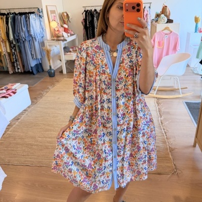 Mulher a tirar selfie em espelho com vestido floral e sandálias pretas, cenário de loja de roupa ao fundo.