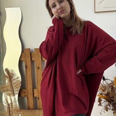 Mulher com camisola vermelha larga e leggings pretas numa sala com espelho ondulado e decoração seca.