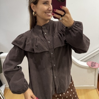 Blusa castanha de veludo com folho largo e botões, com saia castanha às bolinhas, em interior com chão de madeira.