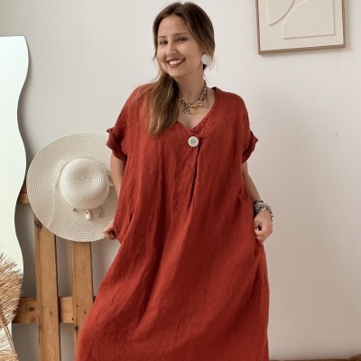 Vestido longo vermelho alaranjado em tecido de linho com botão branco grande no decote em V, usado por mulher sorridente em interior minimalista.