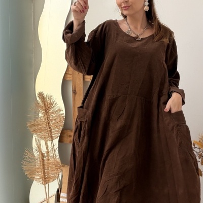 Mulher com vestido castanho escuro de tecido algodão ou linho, em ambiente interior com espelho ondulado e decoração rústica