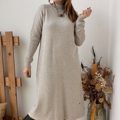 Mulher com vestido cinza claro de malha e botas pretas, com decoração de flores secas num vaso metálico e quadro branco em relevo na parede.