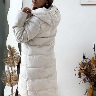 Casaco comprido acolchoado creme com capuz, botas castanhas de pele com bordados, chão de madeira e decoração seca.