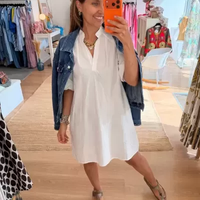 Mulher num vestido branco e casaco denim, em loja de roupa com várias peças penduradas e decoração clara.