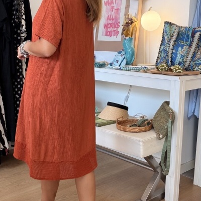 Vestido laranja avermelhado com textura leve e malha na bainha, mulher de cabelo louro, mesa com chapéu, acessórios azuis e verdes e sandálias brancas.