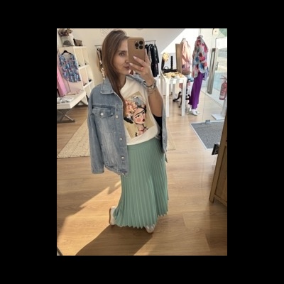 Mulher a experimentar casaco de ganga azul claro, t-shirt branca com estampa e saia plissada verde clara num interior de loja de roupa