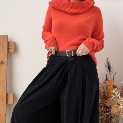 Mulher com camisola laranja de malha e calças largas pretas num ambiente interior com parede branca e flores secas.