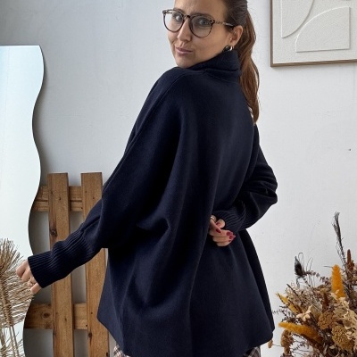 Mulher com camisola preta e calças xadrez num interior decorado com almofada e flores secas.